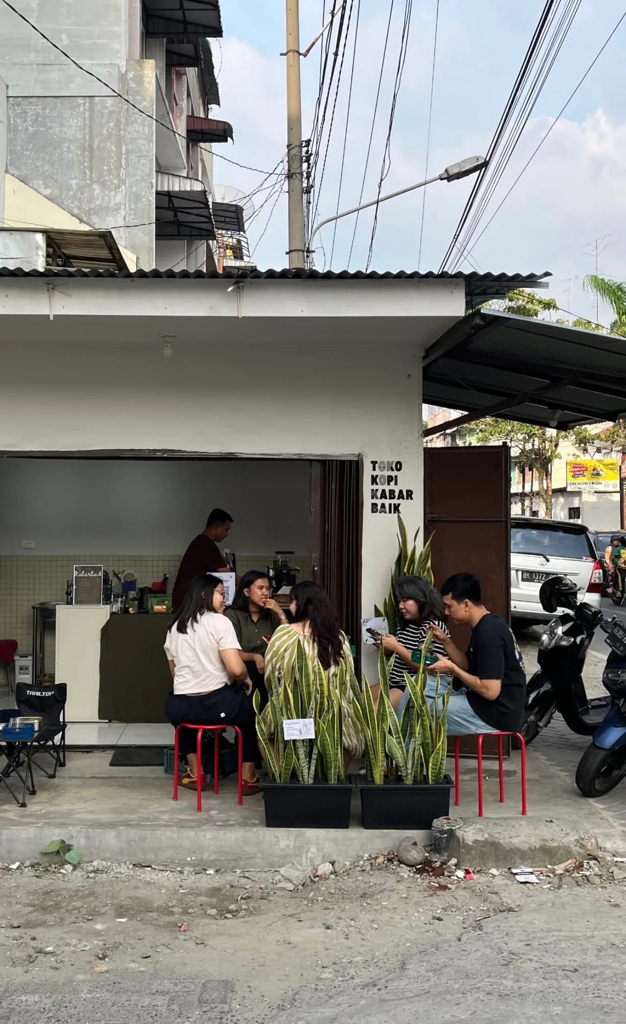 Tamu berkumpul di kursi outdoor Toko Kopi Kabar Baik di siang hari, dengan tanaman lidah mertua sebagai pembatas
